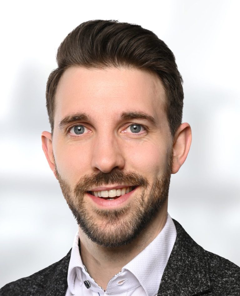 Matthias Palms: lächelnder Mann mit kurzer Frisur, Anzug und hellblauer Bluse vor unscharfem Hintergrund.
