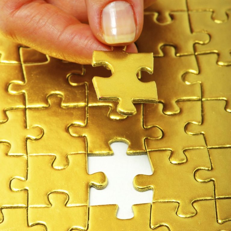 Eine Hand fügt ein goldenes Puzzlestück in ein fast vollständiges Puzzle ein.